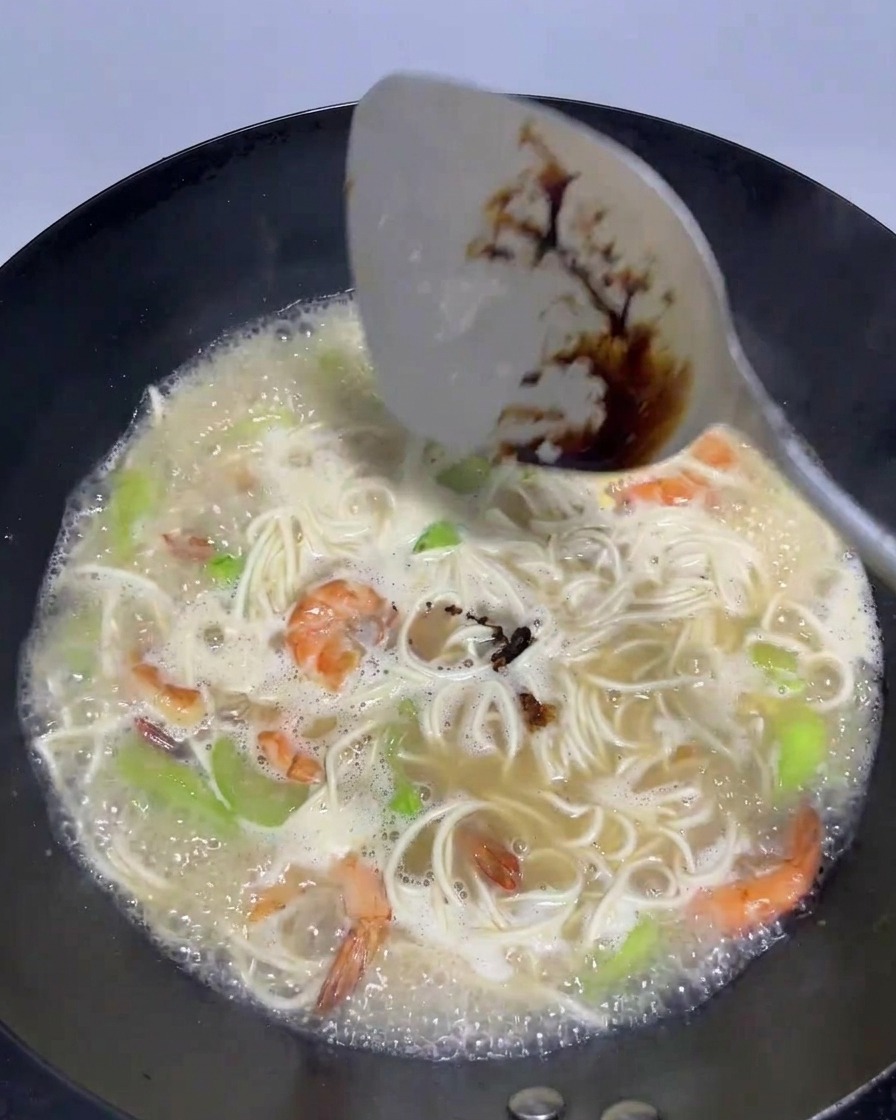 Une spatule versant de la sauce soja foncée dans le bouillon avec les nouilles et les crevettes.