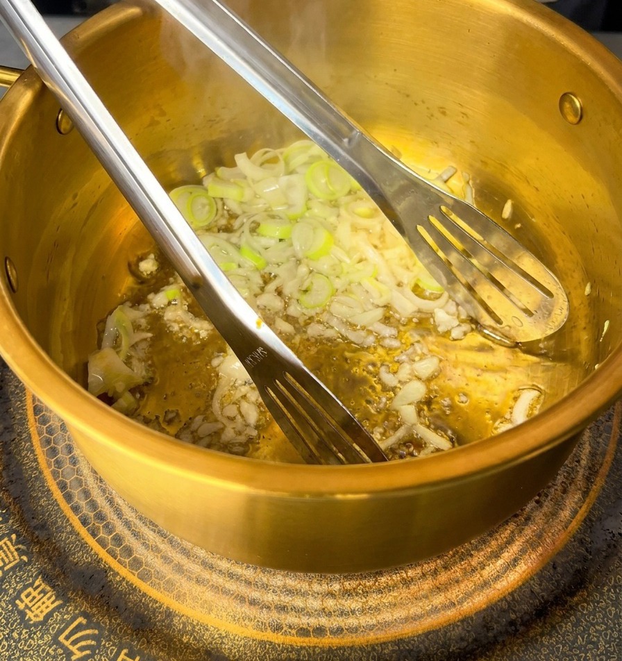 Du porc haché rose mélangé à lail et aux oignons dans la marmite dorée à laide dune pince métallique.