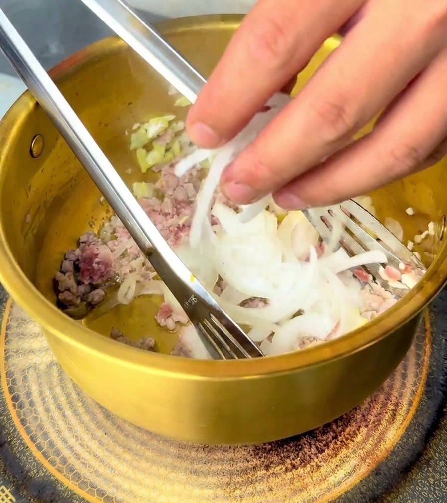 Une main ajoutant une poignée doignons blancs émincés dans la marmite contenant le porc cuit.