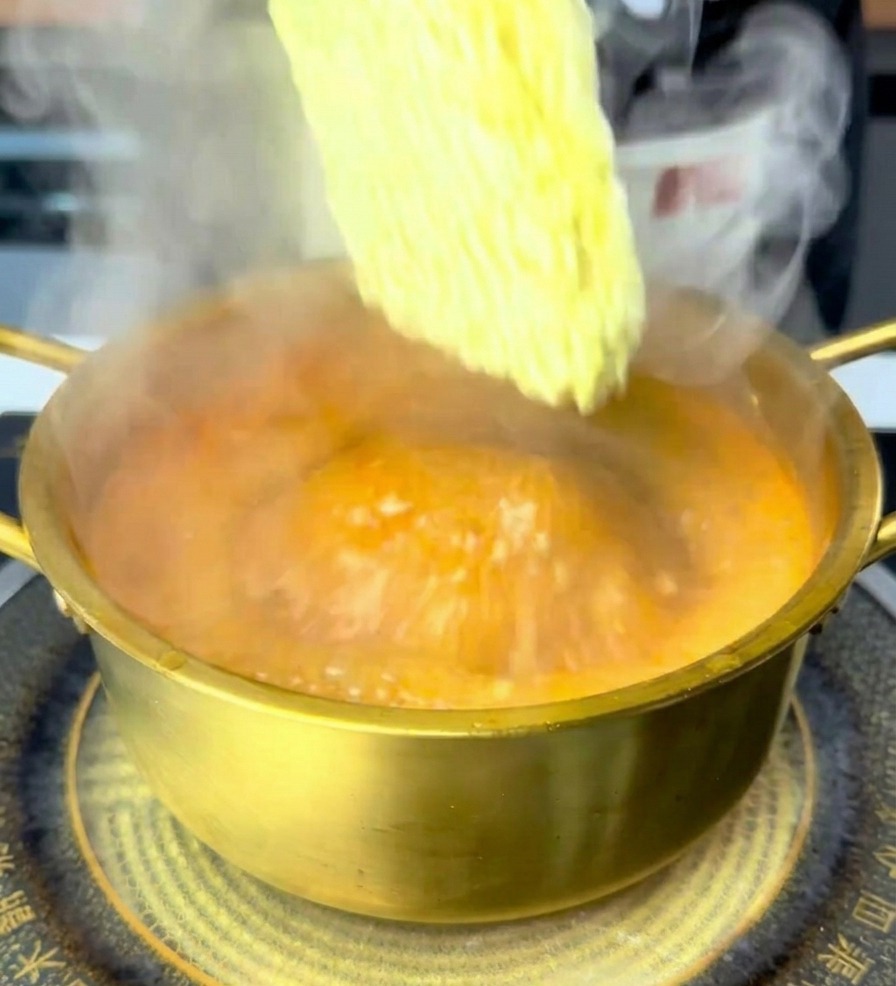 Une main déposant un bloc de nouilles ramen sèches dans le bouillon bouillant rouge orangé.