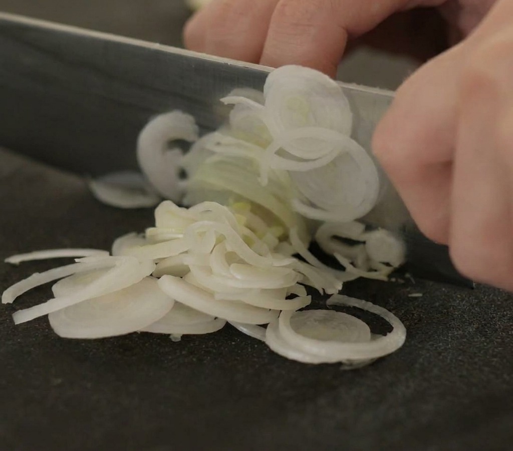 Gros plan de mains utilisant un couteau de chef pour couper des blancs de poireaux en fines rondelles sur une planche noire.