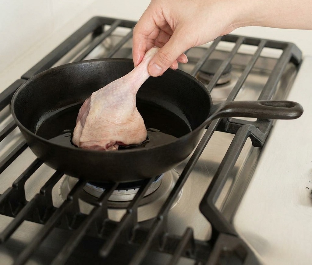 Une main déposant une cuisse de canard crue dans une poêle en fonte chaude sur un brûleur à gaz.