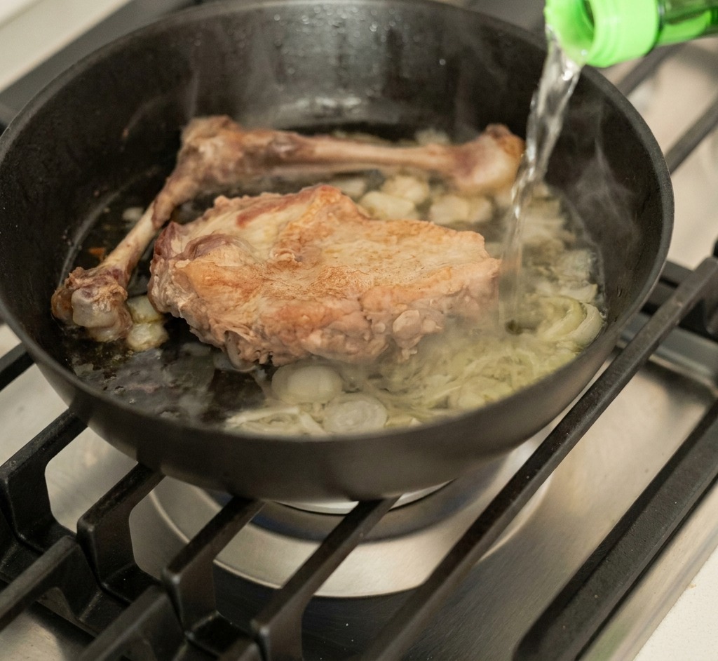 Liquide versé dune bouteille en verre vert dans une poêle fumante contenant le canard et les aromates.