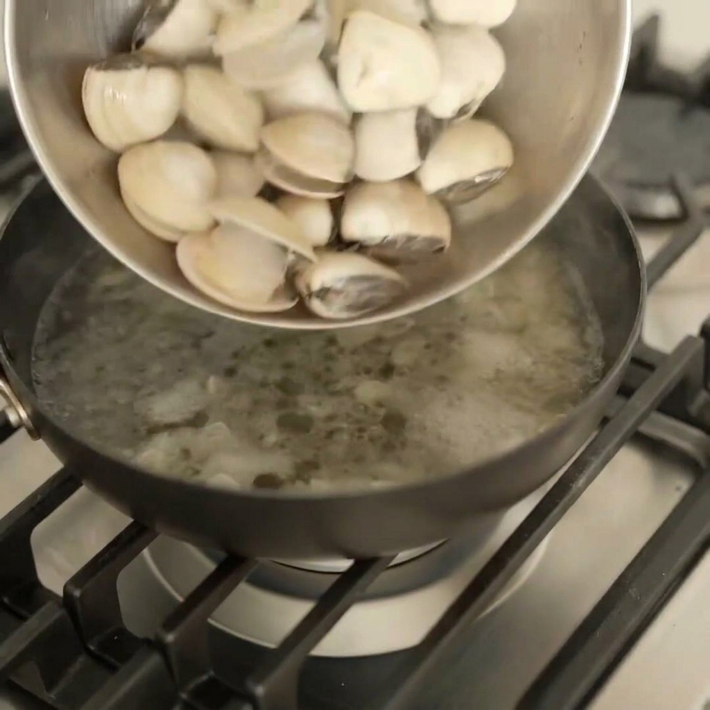 Palourdes blanches fraîches versées dun bol en métal dans une poêle sombre sur une cuisinière à gaz.