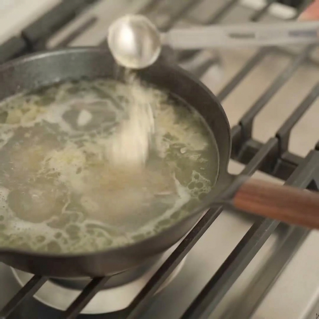 Une cuillère doseuse en argent versant un liquide sombre dans une casserole de bouillon de ramen clair.