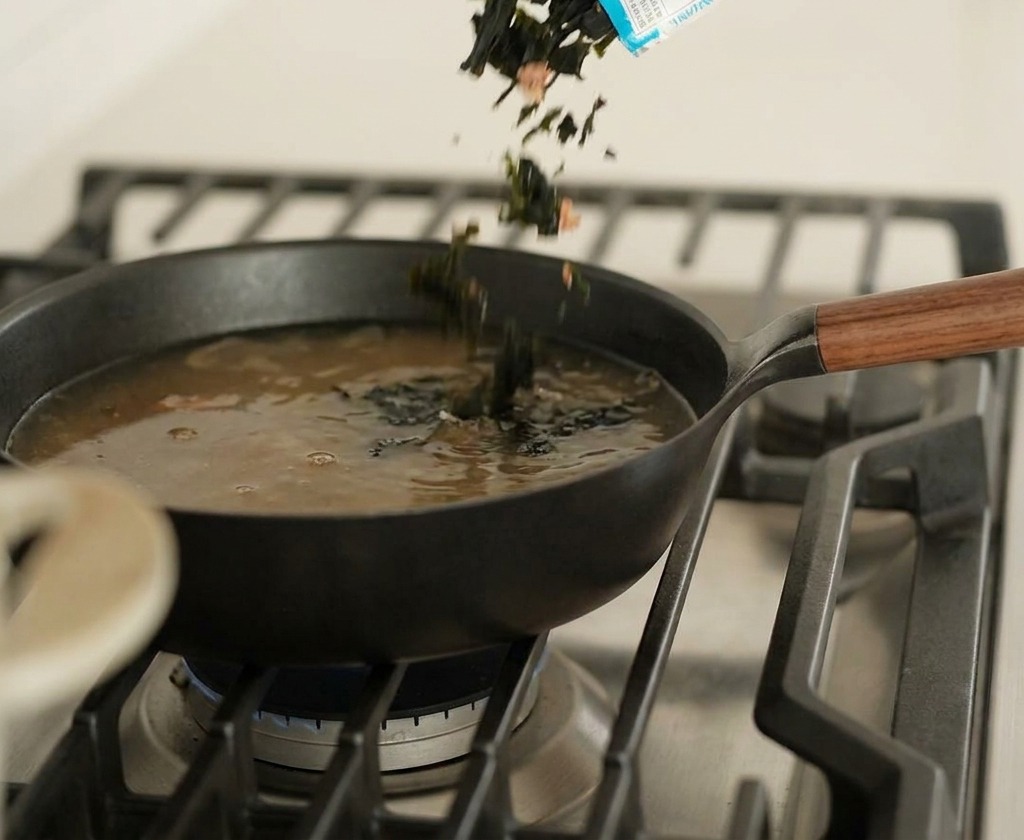 Morceaux dalgues séchées versés dun emballage bleu et blanc dans une poêle contenant un bouillon sombre.