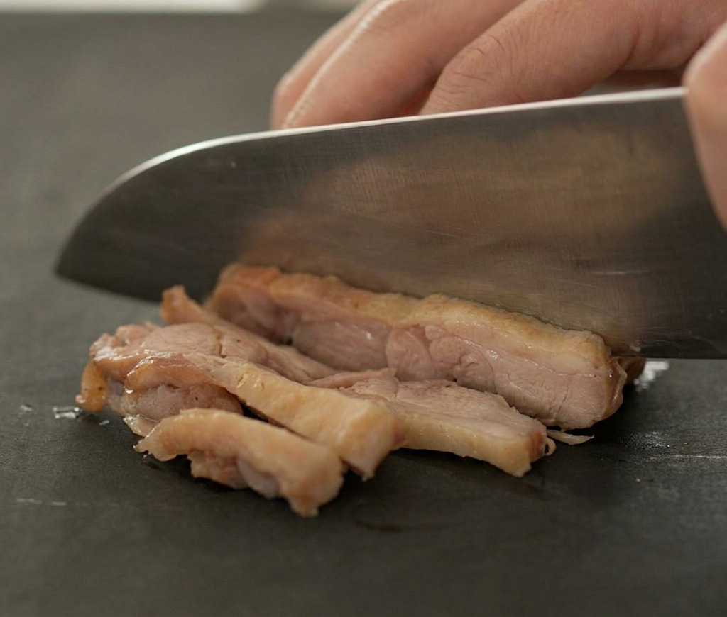 Un couteau tranchant coupant un morceau de canard doré en fines lamelles sur une planche sombre.