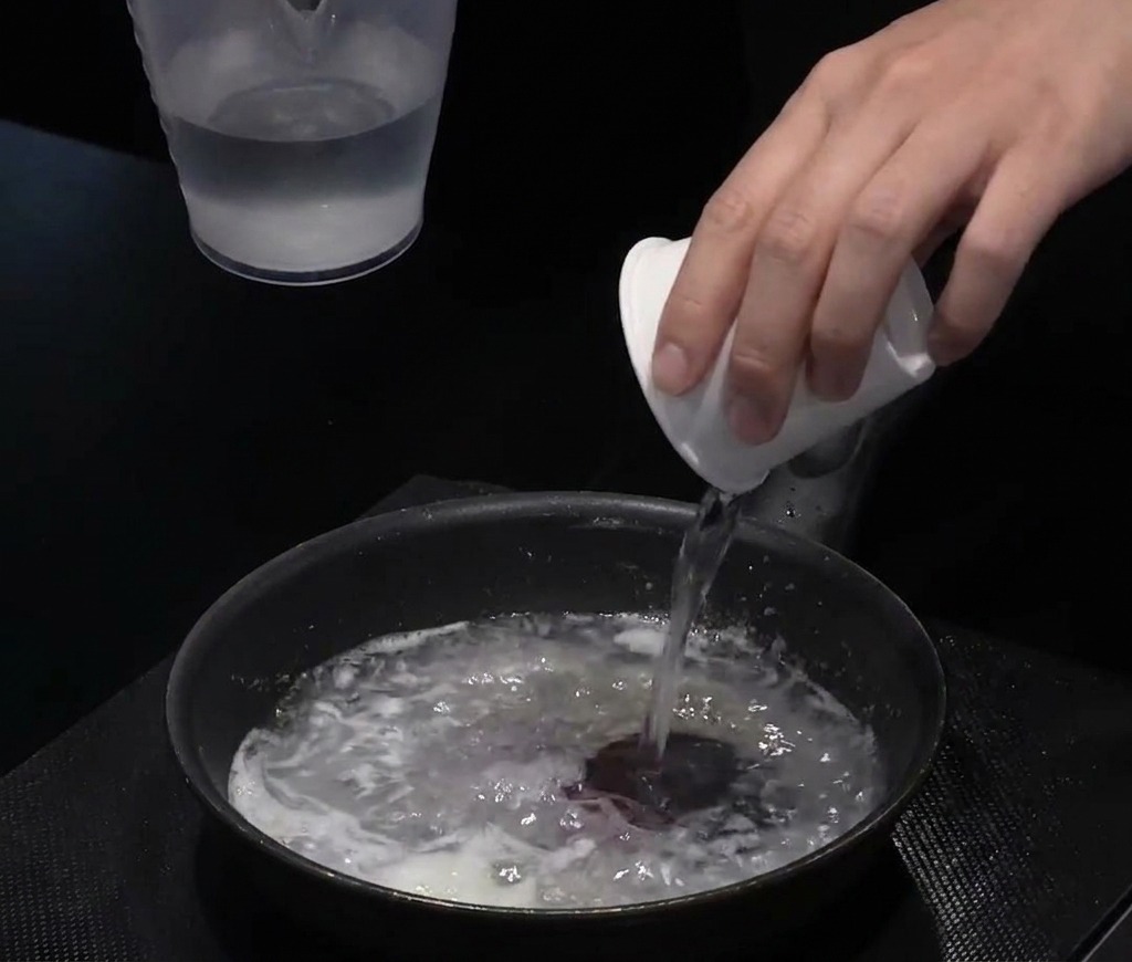 De leau versée dune petite tasse blanche dans une poêle noire contenant un mélange dhuile et de farine.