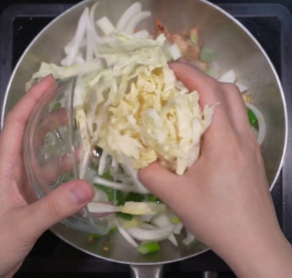 Des mains ajoutant une large portion de chou chinois râpé dans une poêle contenant des oignons sautés.