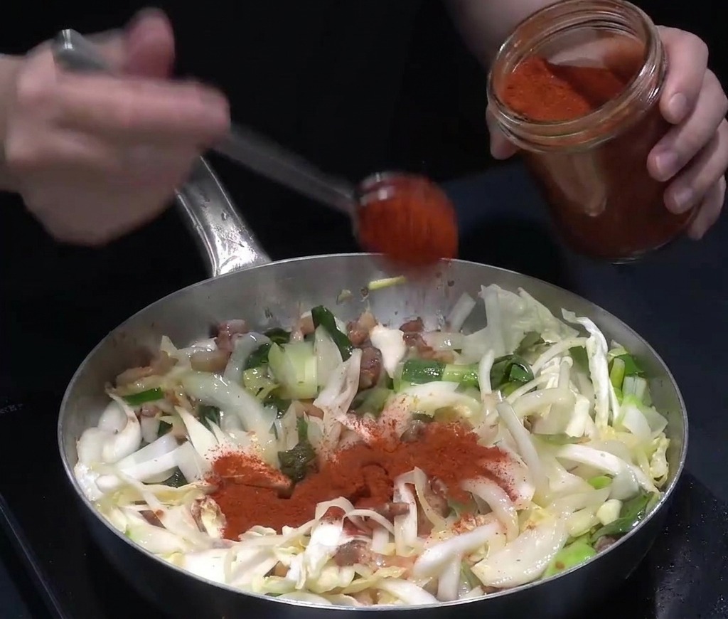 Poudre de piment rouge vif versée dun pot en verre sur un mélange de légumes et de porc sautés.