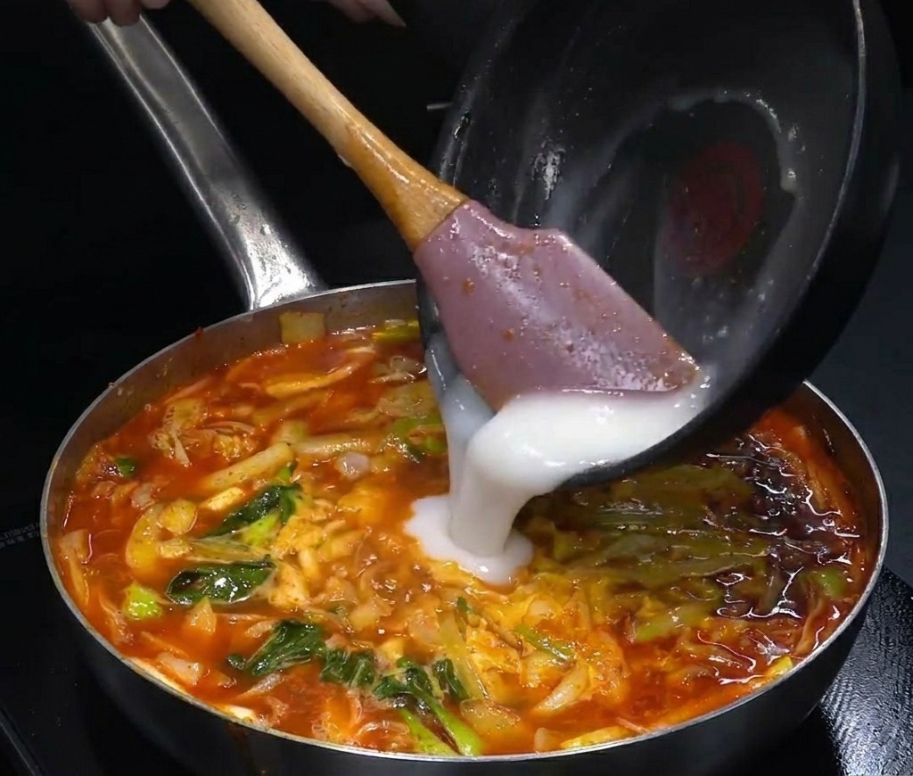 Un liquide blanc opaque versé dune petite poêle noire dans une grande marmite de soupe rouge bouillonnante.
