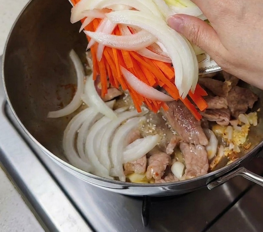 Une main versant une assiette doignons émincés, de carottes et de courgettes dans la marmite avec la viande.