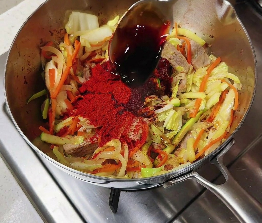 Une cuillère en argent versant de la sauce soja foncée sur les légumes sautés couverts de piment rouge dans une marmite en inox.