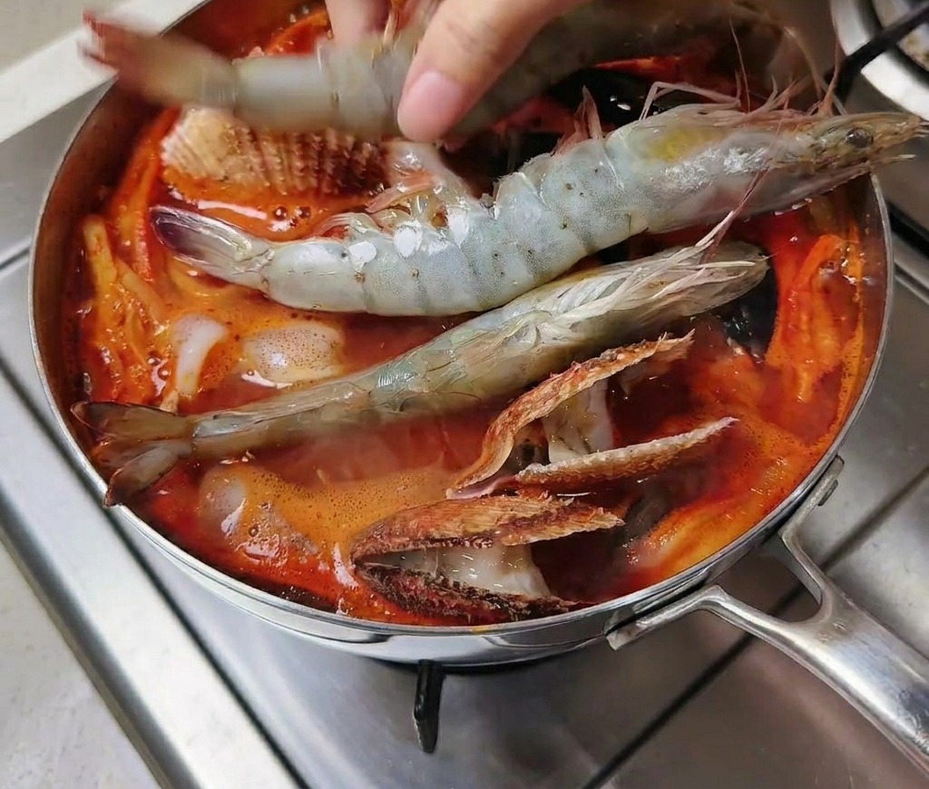 Une main déposant plusieurs grosses crevettes grises crues dans une marmite de soupe rouge épicée remplie de coquillages.
