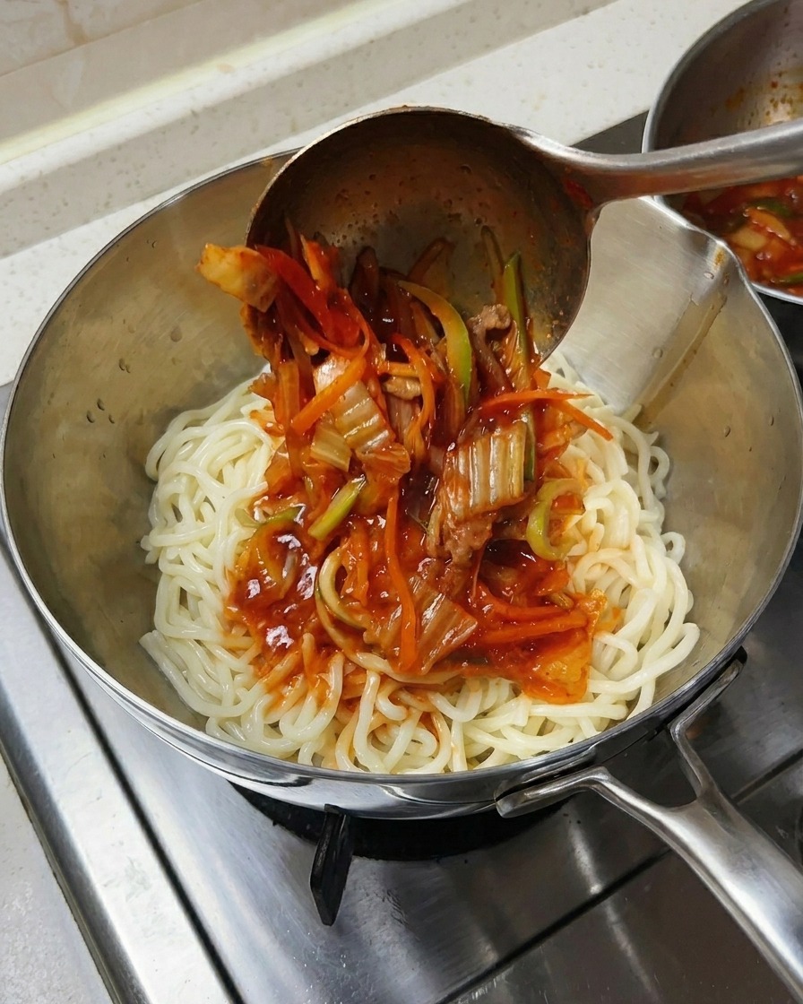 Une louche métallique versant de la soupe rouge épicée avec fruits de mer et légumes sur un dôme de nouilles blanches dans un bol en inox.