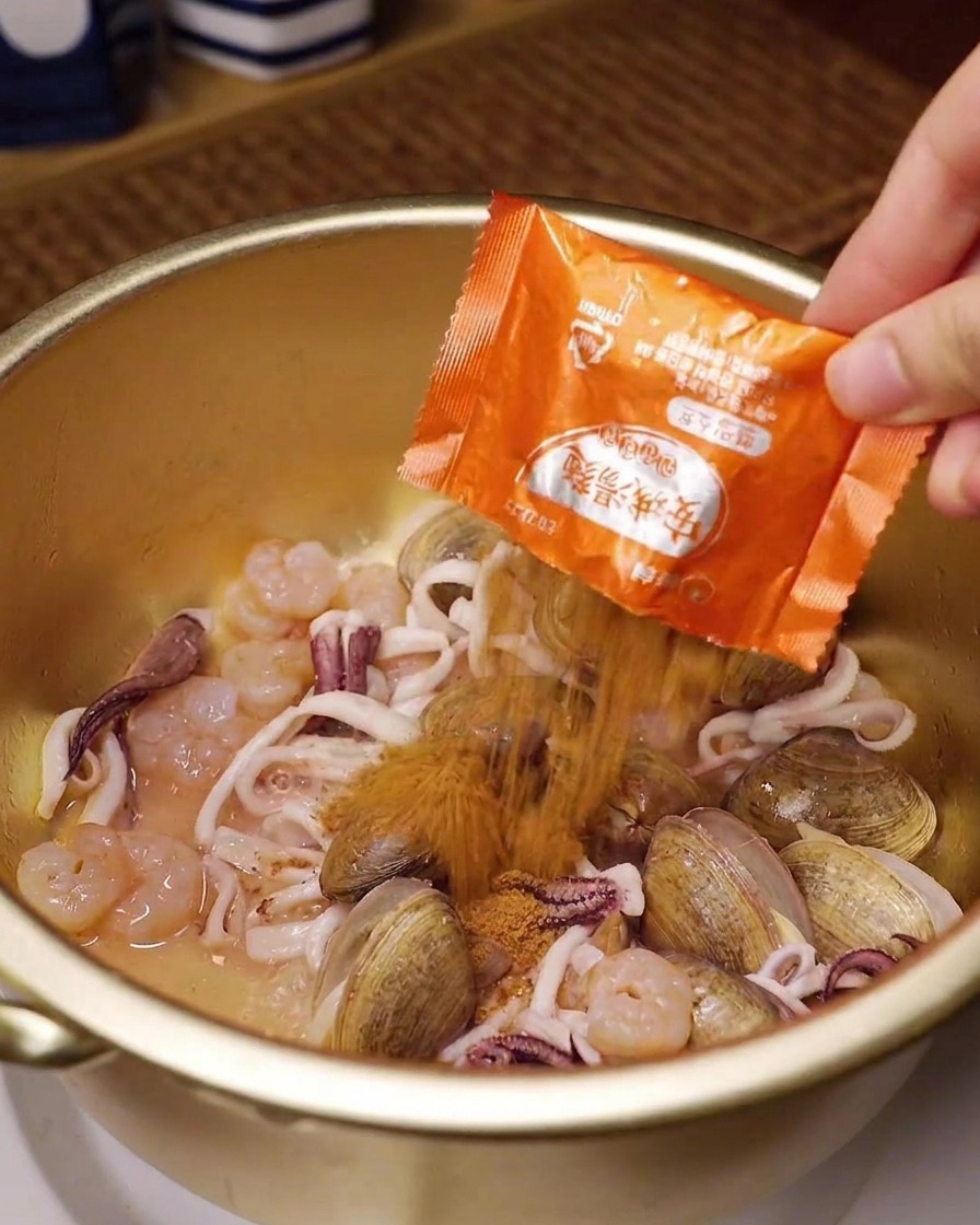 Une main versant un sachet orange dassaisonnement pour ramen instantané sur des palourdes, des calmars et des crevettes dans une marmite en laiton.