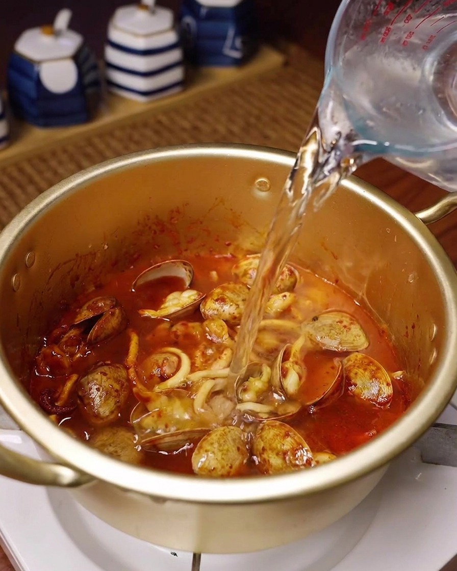 Eau fraîche versée dun pichet doseur en verre dans une marmite dorée contenant la base de fruits de mer épicée.