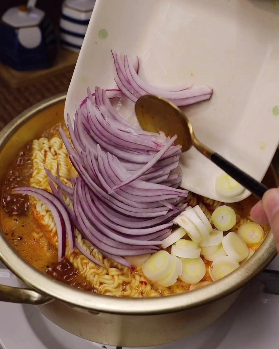 Tranches fines doignons rouges et de cébettes blanches ajoutées dans une marmite de ramen frémissant dans un bouillon rouge.