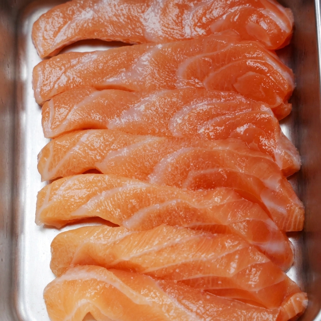 Tranches de saumon frais disposées dans un plateau métallique, saupoudrées de sel fin et de grains de poivre noir.