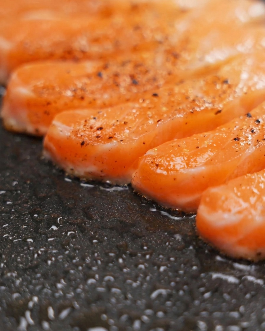 Filets de saumon assaisonnés grésillant dans une poêle antiadhésive sombre, montrant une coloration dorée et des grains de poivre en surface.