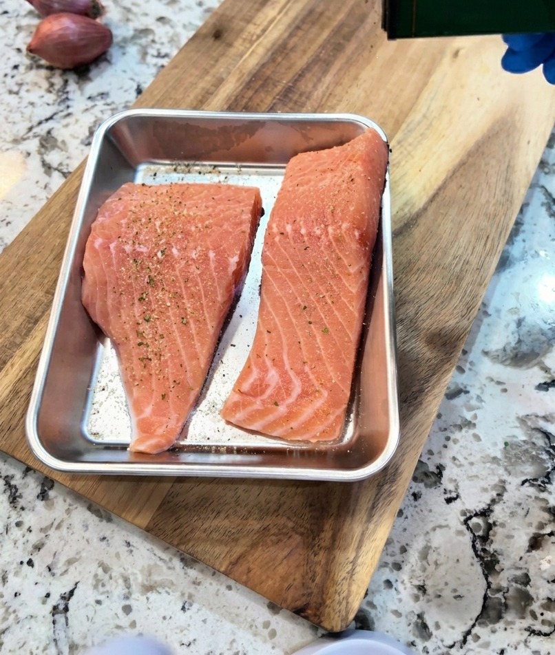 Une main assaisonnant des filets de saumon crus dans un plateau métallique avec du poivre noir et de lail.
