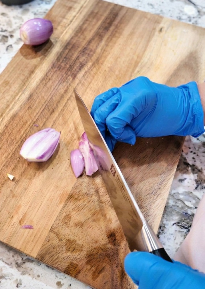 Un couteau de cuisine coupant des échalotes violettes en fines rondelles sur une planche en bois.