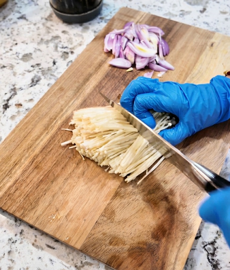 Un couteau coupant la base dun bouquet de champignons enoki blancs sur une planche à découper.