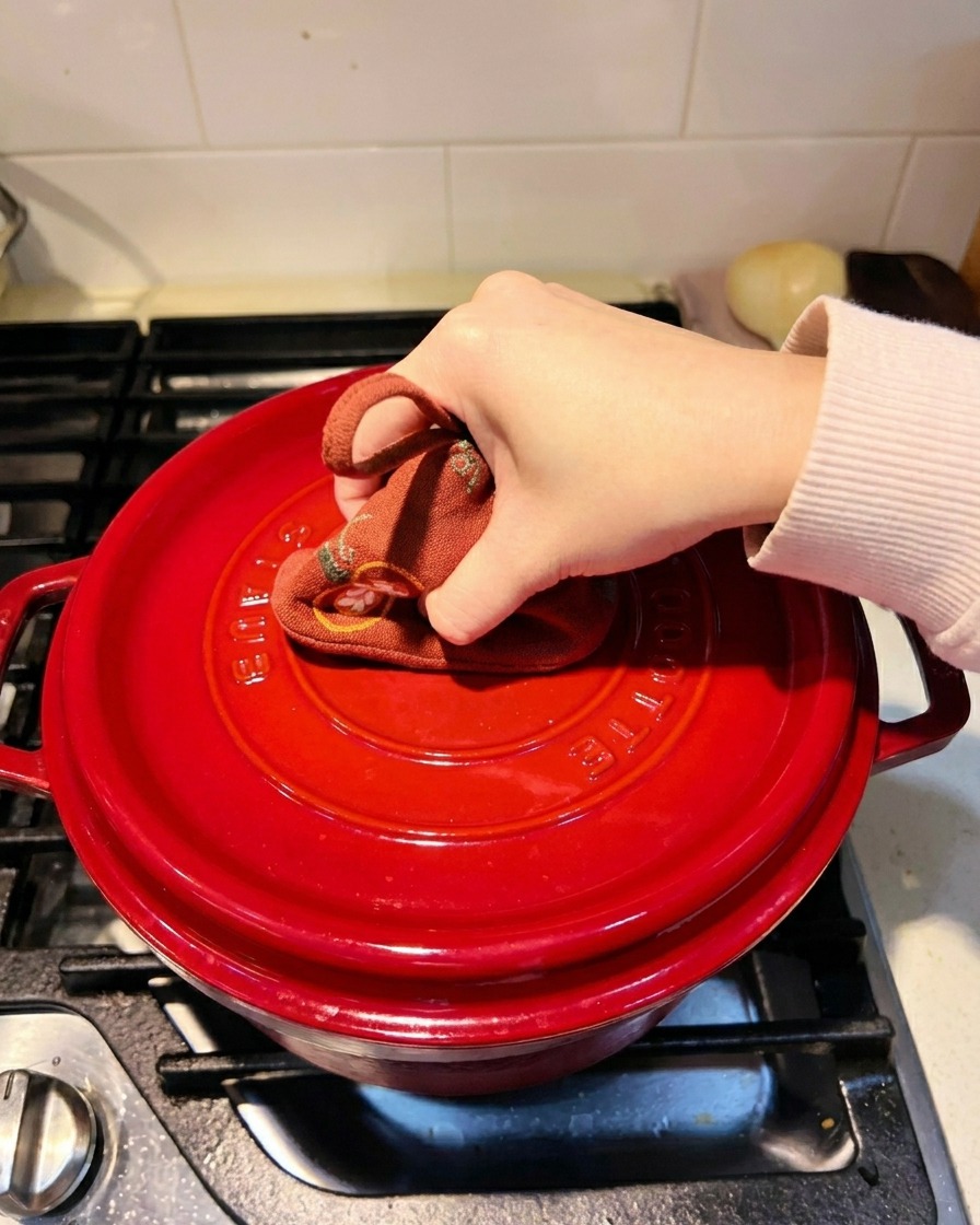 Une main utilisant une manique pour presser le couvercle dune cocotte rouge sur une cuisinière.
