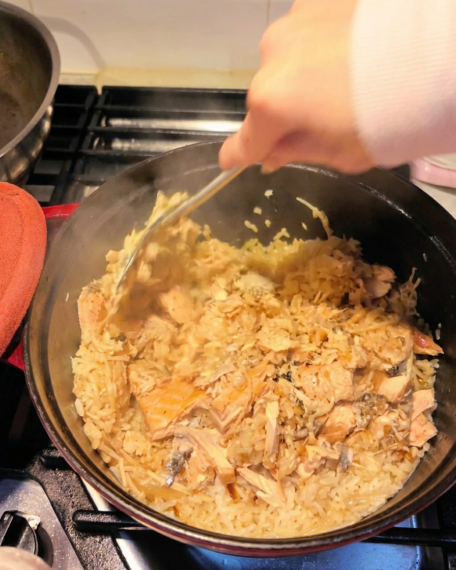 Une main utilisant une spatule pour défaire les filets de saumon et les mélanger au riz assaisonné.