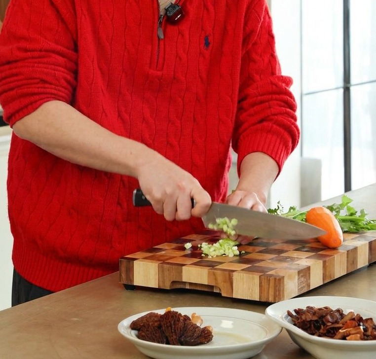 Une personne utilisant un couteau professionnel pour couper finement du céleri vert et des carottes oranges sur une planche en bois.