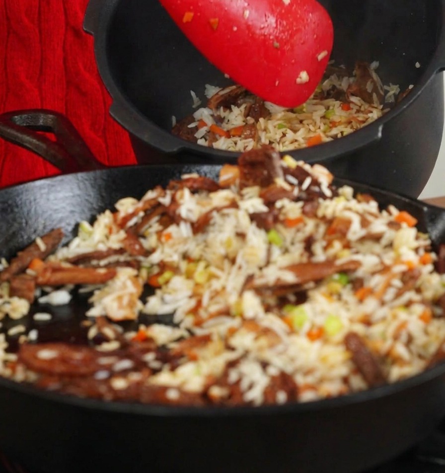 Spatule rouge transférant le riz sauté et les champignons dune poêle vers la cuve dun cuiseur à riz noir.