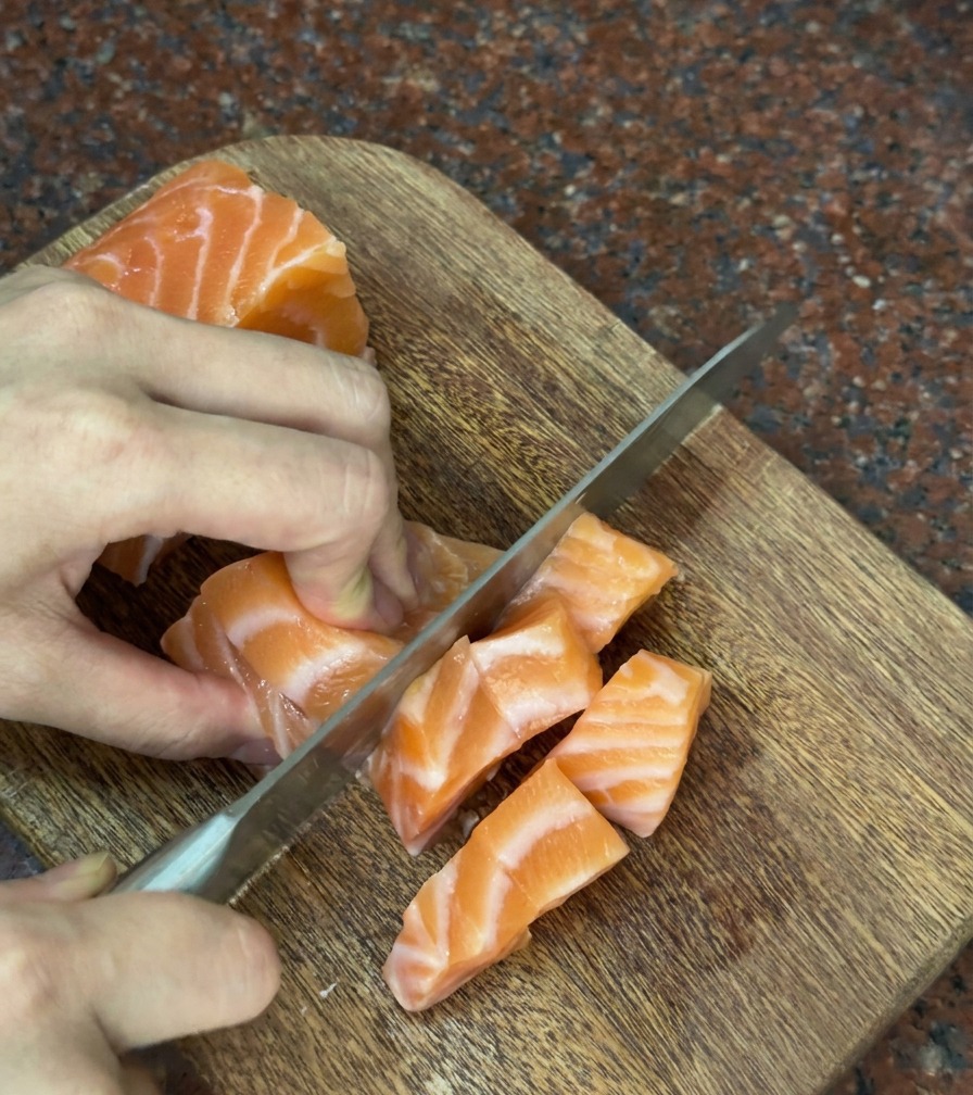 Mains coupant soigneusement un morceau de saumon frais en cubes réguliers sur une planche en bois.