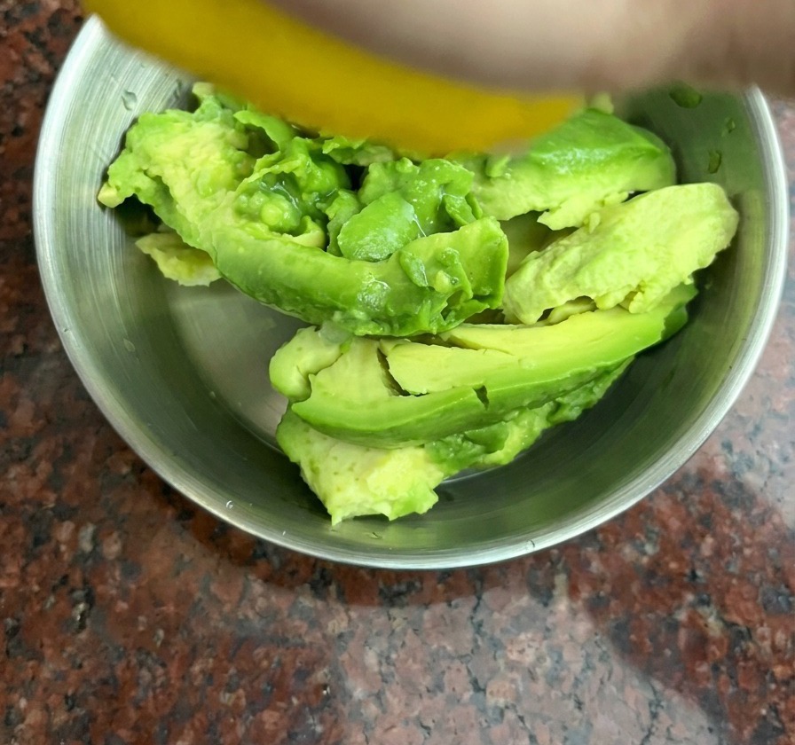 Une main pressant un citron jaune sur des morceaux davocat dans un bol en inox.