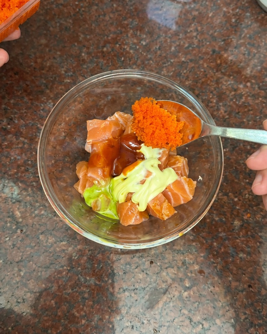 Une cuillère ajoutant une portion généreuse de petits œufs de poisson orange dans le bol de saumon.