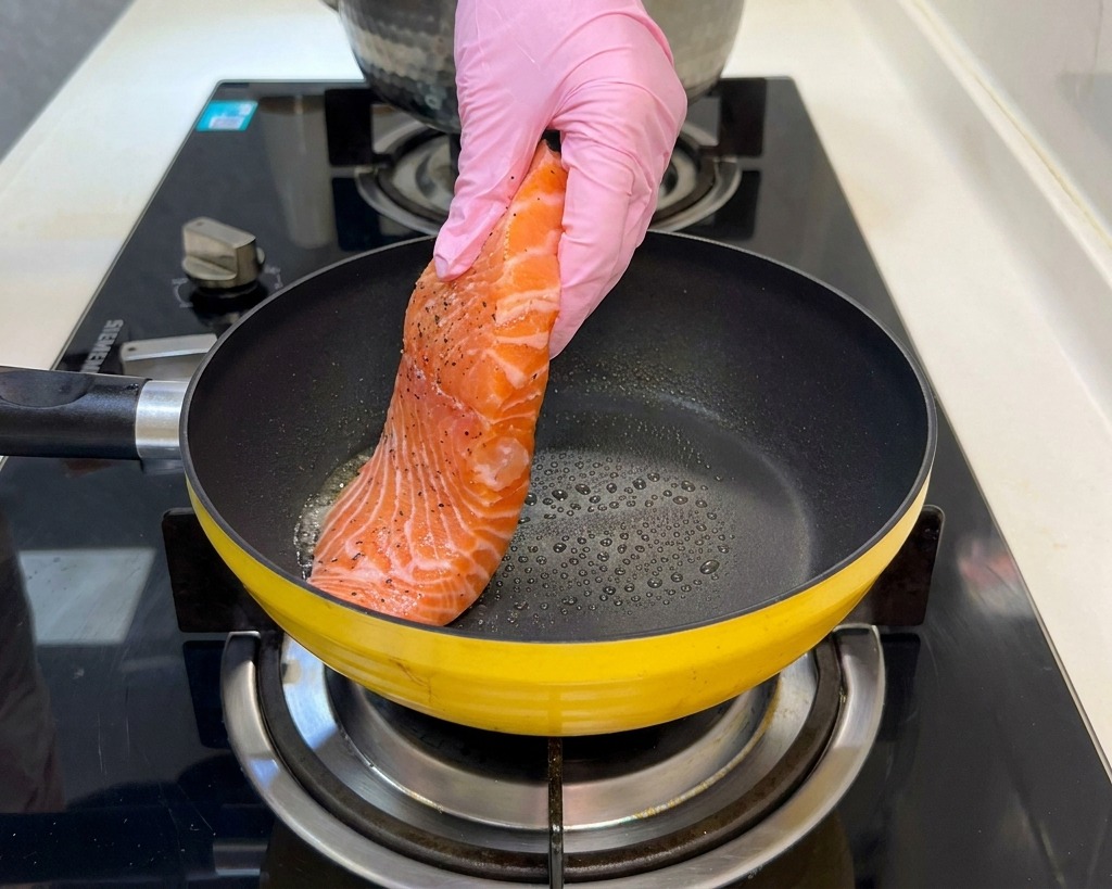 Un filet de saumon cru assaisonné est déposé dans une poêle jaune chauffée par une main gantée de rose.