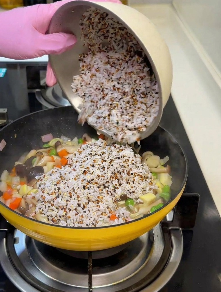 Une main gantée de rose verse un bol de riz blanc et de quinoa mélangés dans une poêle contenant des légumes sautés.