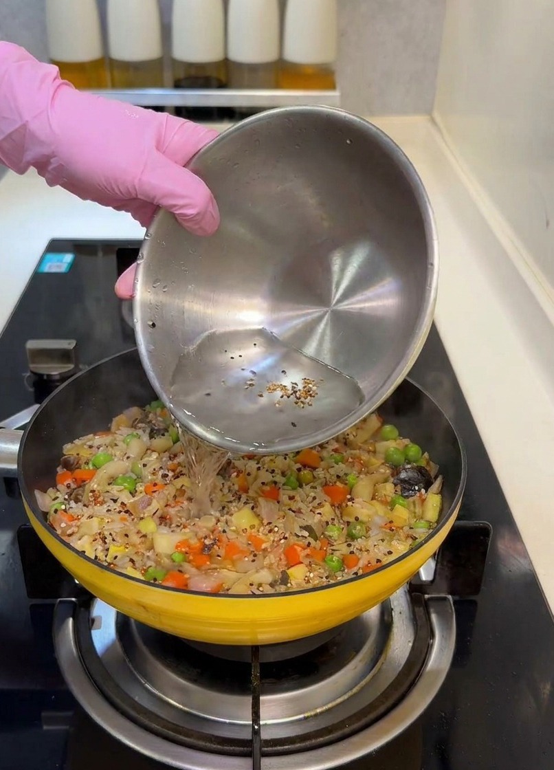 De leau versée dun bol en inox dans une poêle jaune remplie dun mélange coloré de riz et de légumes.