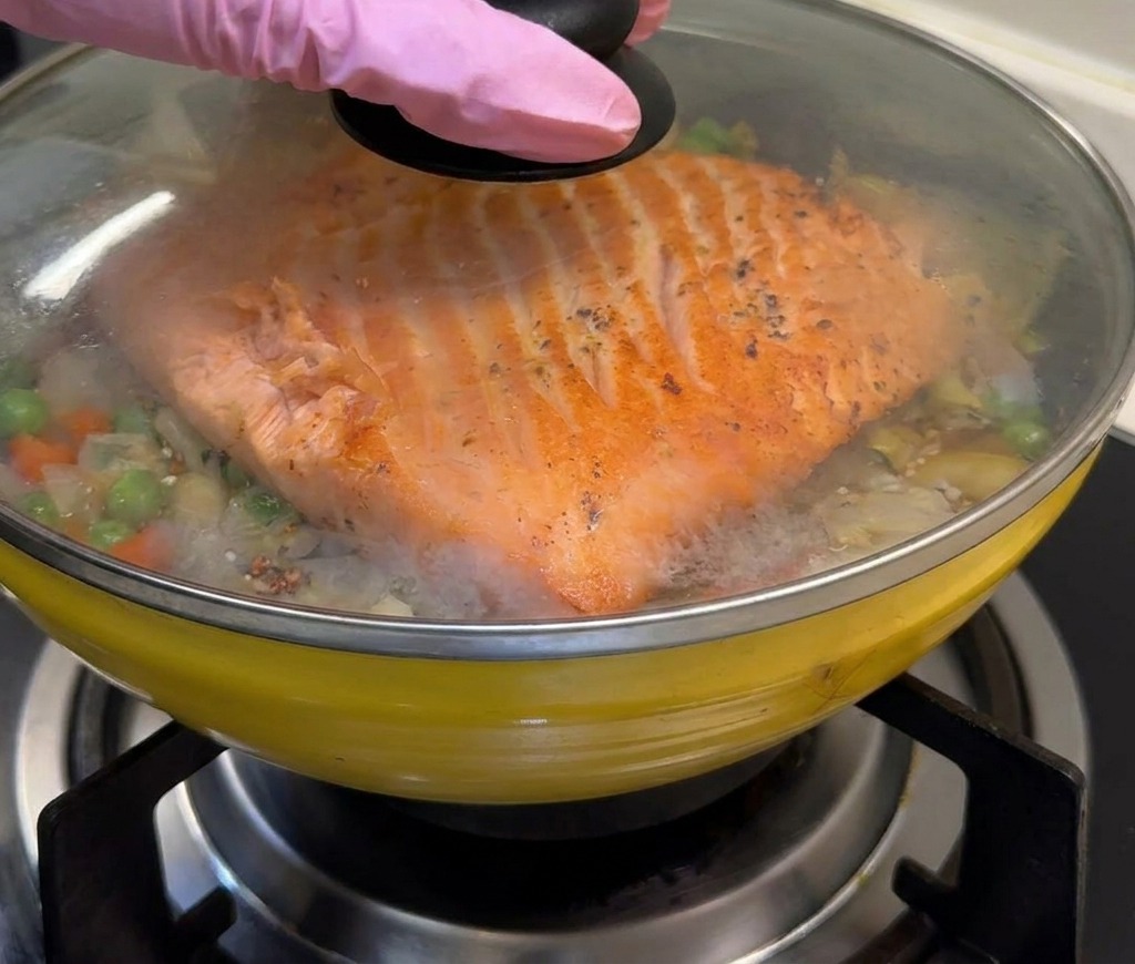 Une poêle jaune contenant le saumon et le riz aux légumes est couverte dun couvercle en verre pour emprisonner la vapeur.
