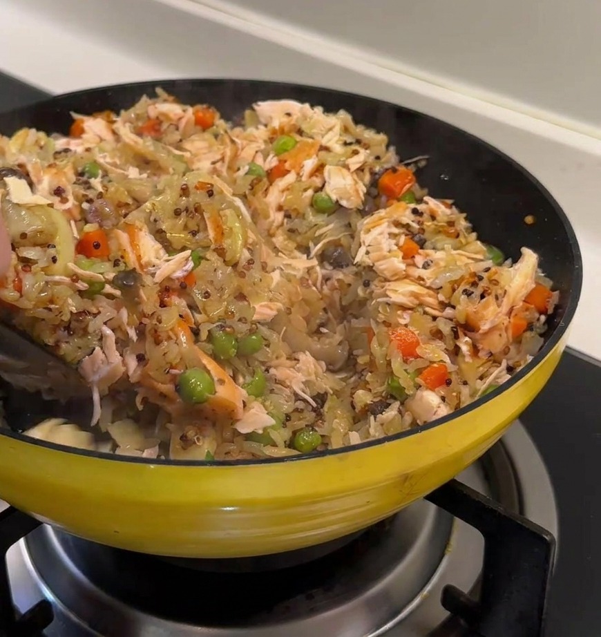 Une main utilisant une spatule noire pour incorporer de larges morceaux de saumon dans un riz coloré au quinoa, pois et carottes.