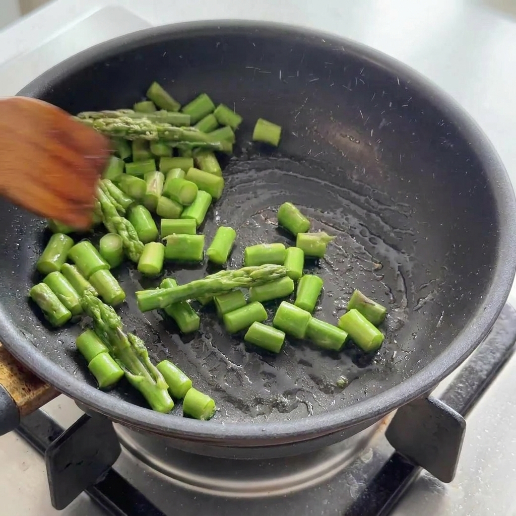 Des morceaux dasperges vertes sautés dans une poêle noire avec une spatule en bois.