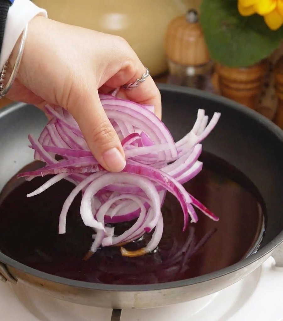 Une main jetant des oignons rouges émincés dans une poêle remplie de sauce sombre frémissante.