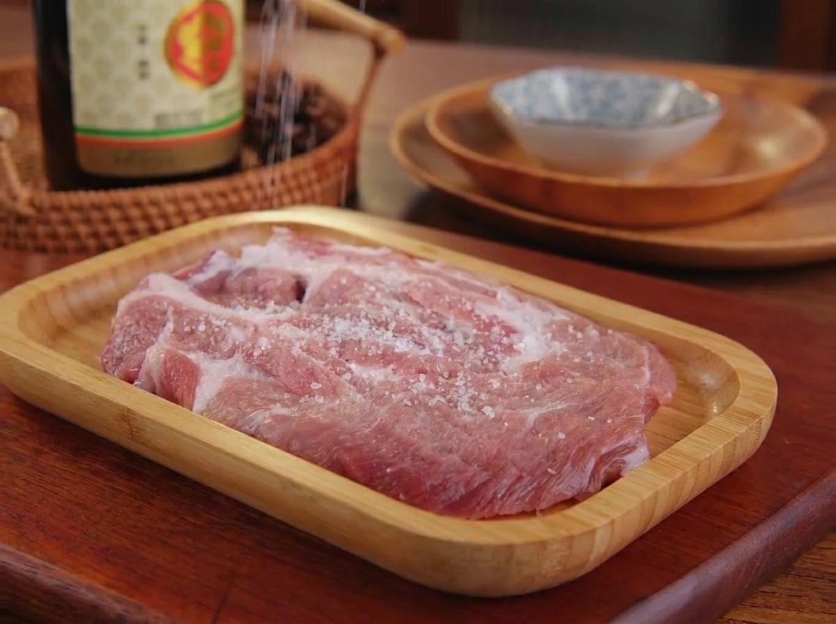 Du sel fin et du poivre noir saupoudrés sur une pièce de porc crue posée sur un plateau en bois.
