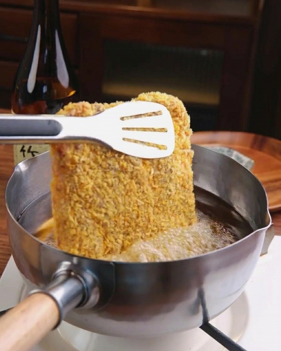 Une escalope de porc panée plongée dans une casserole en inox remplie dhuile de friture chaude et bouillonnante.