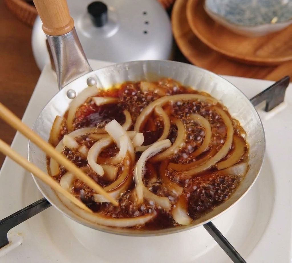 Des oignons blancs finement tranchés mijotant dans une sauce brune à lintérieur dune petite poêle sur un réchaud à gaz.