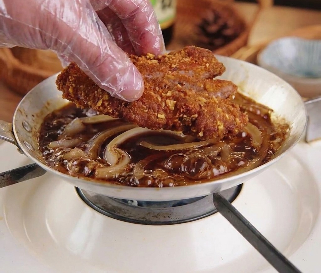 Une main gantée plaçant une escalope de porc frite et tranchée dans une poêle doignons frémissants.