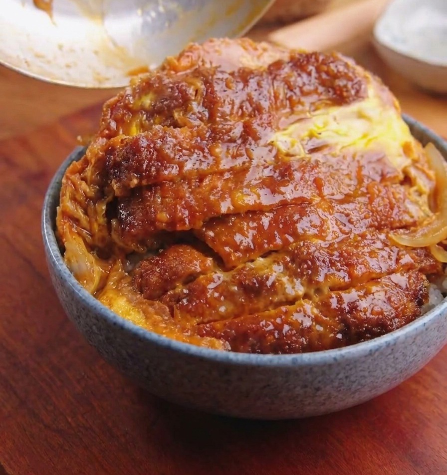 Une poêle inclinée au-dessus dun bol de riz pour faire glisser le porc pané aux œufs et oignons sur le riz.