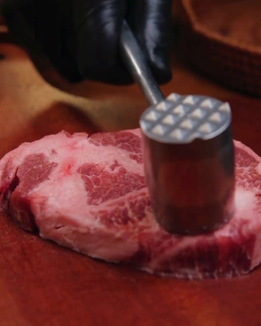 Main gantée utilisant un marteau à viande en métal pour attendrir une épaisse tranche déchine de porc crue sur une planche en bois.
