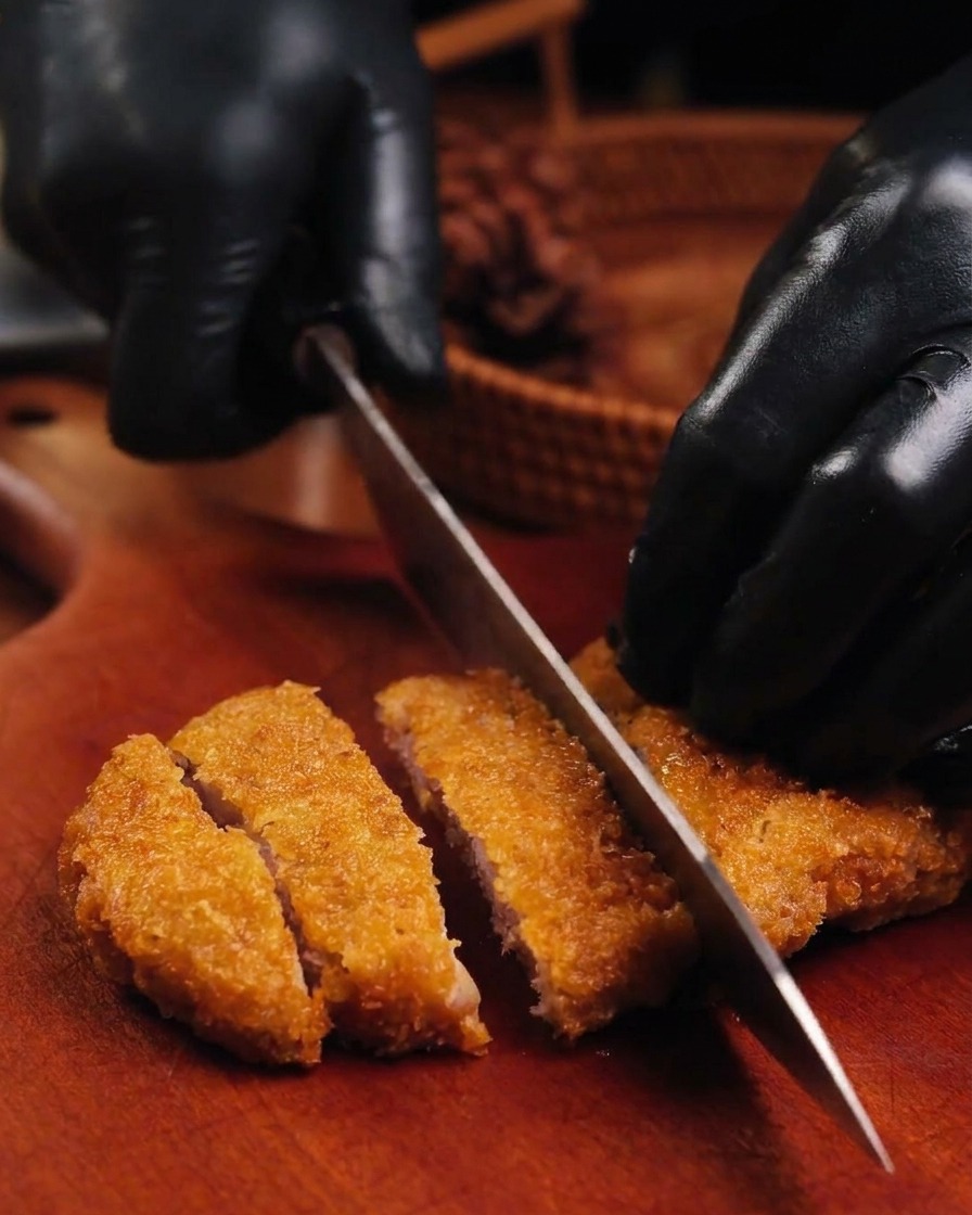 Couteau tranchant une escalope de porc frite bien dorée sur une planche en bois tenue par des mains gantées.