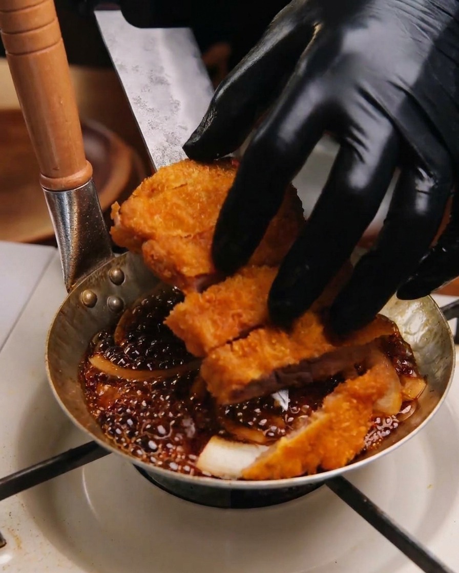 Mains gantées disposant les tranches de porc frit dans une poêle sur un lit doignons blancs et de sauce sombre.