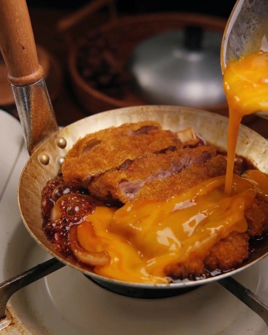 Mélange dœufs crus battus versé dun bol sur une escalope de porc mijotant dans la sauce avec des oignons.